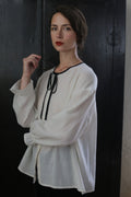 A woman with dark hair, styled in a loose updo, stands against a dark background. She wears a Ribbon Shirt - Japanese Wool Gauze with a black ribbon tie at the neckline. Her expression is serene, and she has red lipstick on. One hand is raised near her face.