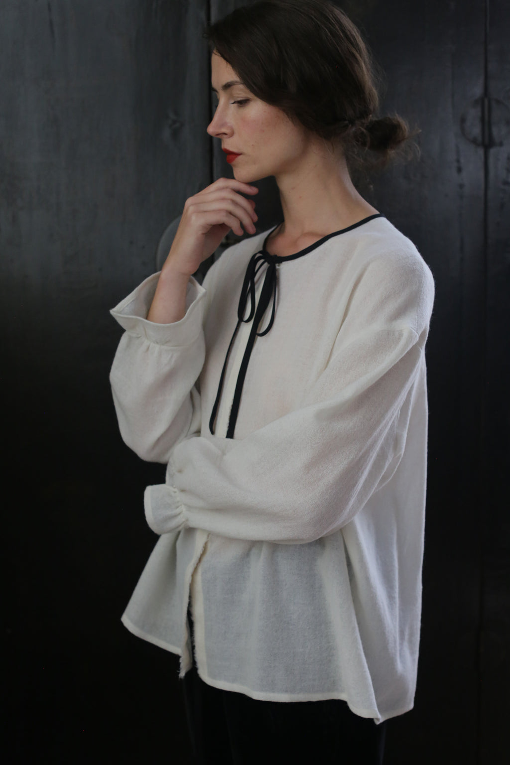 A woman with brown hair tied in a loose bun is seen in profile against a dark background. She is wearing a loose-fitting, long-sleeved Ribbon Shirt - Japanese Wool Gauze with a black ribbon tie at the neckline. Her right hand is gently touching her chin, and she appears thoughtful.