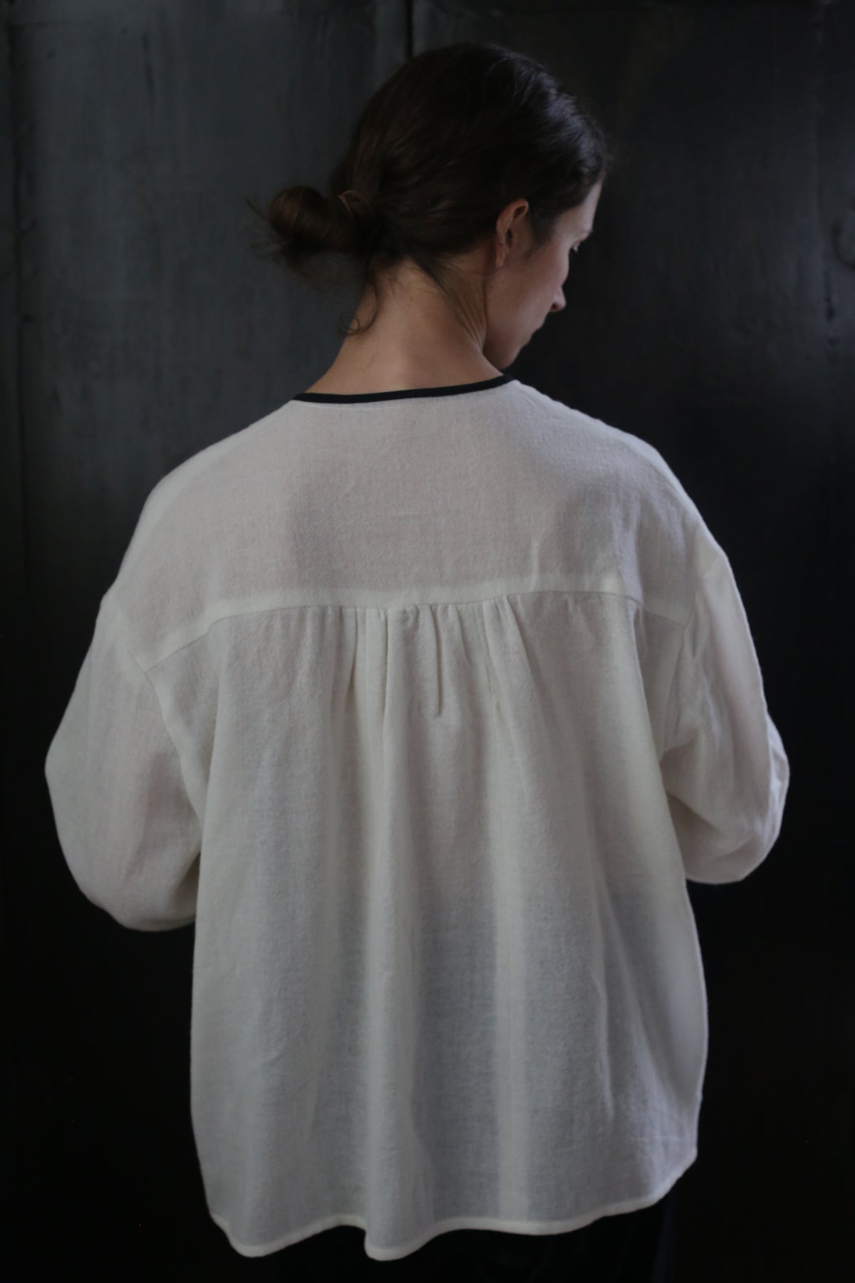 A person with dark hair tied in a low bun stands with their back to the camera, wearing a loose-fitting, long-sleeved white Ribbon Shirt - Japanese Wool Gauze. The background is a dark, solid color.