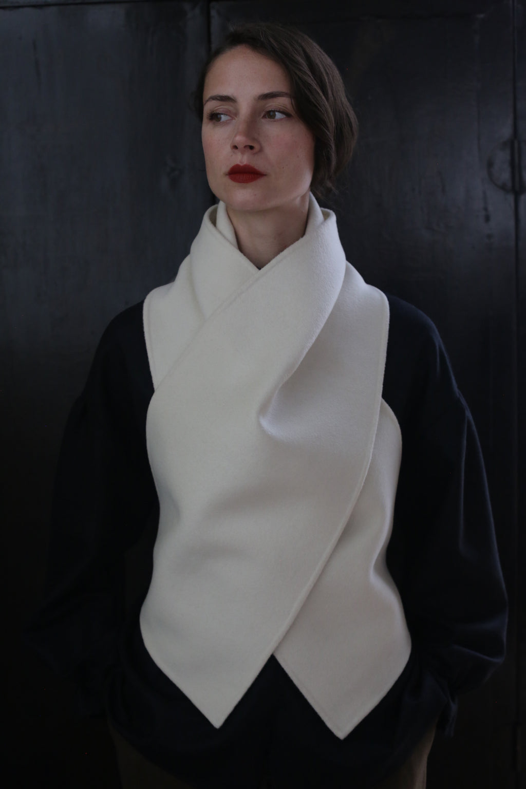 A person poses against a dark background, wearing a black top and a cream-colored, scarf-like garment draped around the neck and chest in a cross-over fashion. The X Scarf - Double-face Cashmere | Undyed features ceramic buttons as accents. They have short, straight hair and wear red lipstick, looking slightly off to the side.