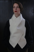 A person poses against a dark background, wearing a black top and a cream-colored, scarf-like garment draped around the neck and chest in a cross-over fashion. The X Scarf - Double-face Cashmere | Undyed features ceramic buttons as accents. They have short, straight hair and wear red lipstick, looking slightly off to the side.