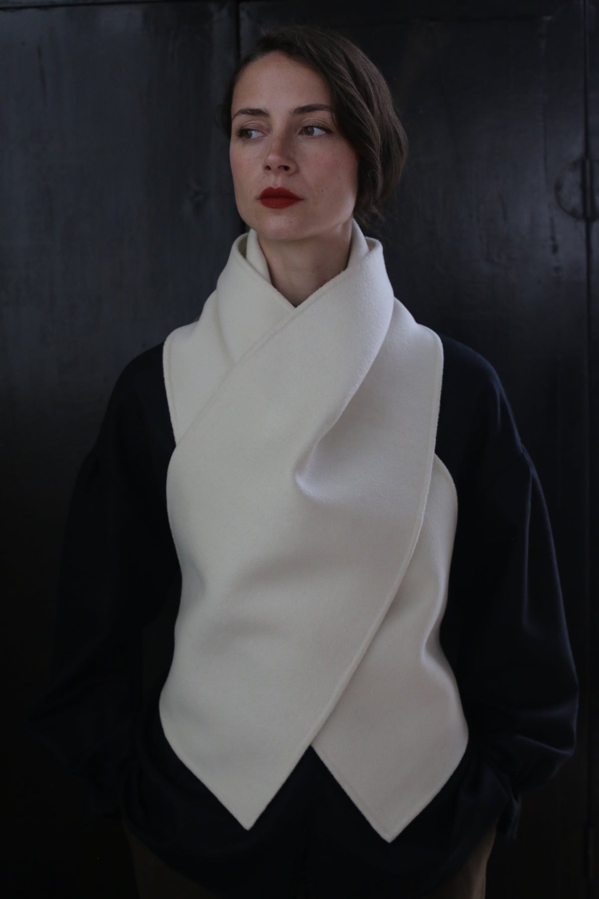A person poses against a dark background, wearing a black top and a cream-colored, scarf-like garment draped around the neck and chest in a cross-over fashion. The X Scarf - Double-face Cashmere | Undyed features ceramic buttons as accents. They have short, straight hair and wear red lipstick, looking slightly off to the side.