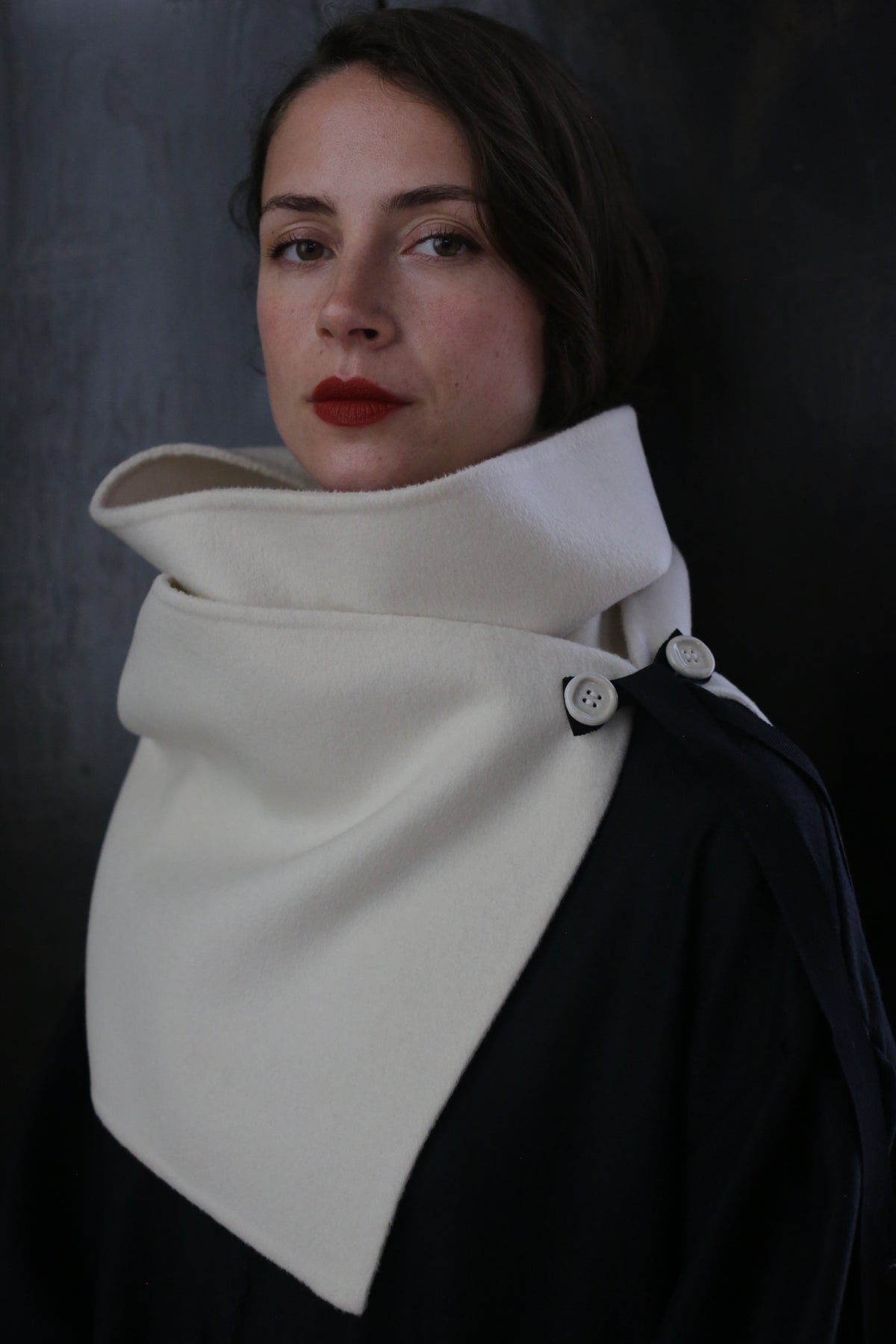A woman with dark hair and red lipstick is wearing a stylish ivory X Scarf - Double-face Cashmere | Undyed, accented with ceramic buttons, against a dark background. The scarf is wrapped around her neck and shoulders, creating a sophisticated and elegant look.