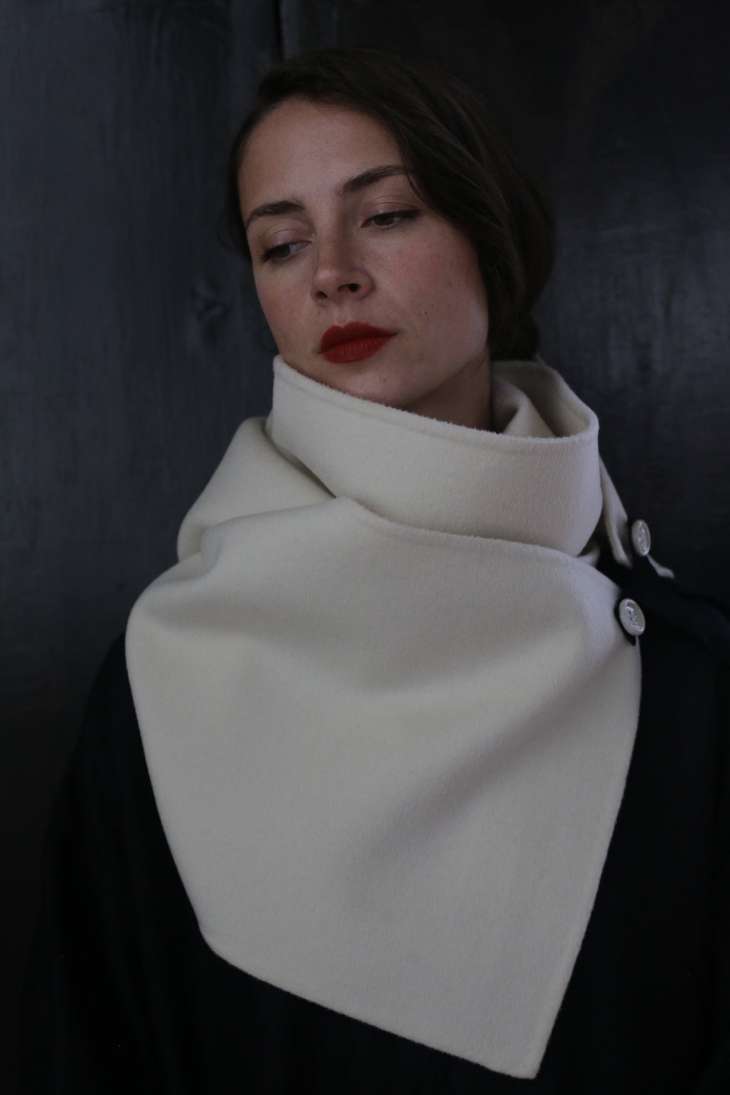 A person with long dark hair and red lipstick wears a fashionable black coat made of double-faced cashmere, adorned with ceramic buttons and a large, high-collared white X Scarf - Double-face Cashmere | Undyed buttoned at the side. They look downward with a solemn expression, standing against a dark background.