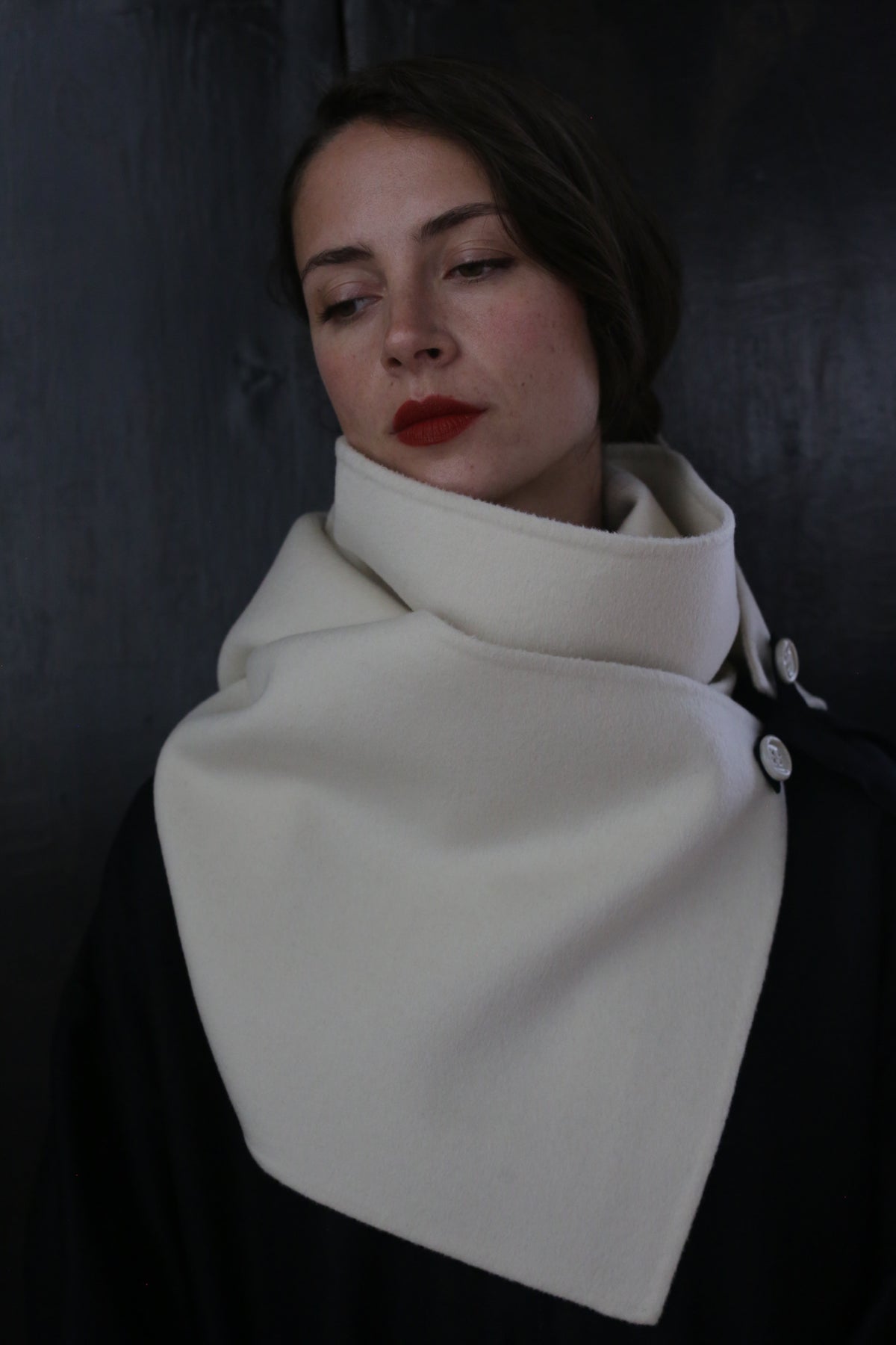 A person with long dark hair and red lipstick wears a fashionable black coat made of double-faced cashmere, adorned with ceramic buttons and a large, high-collared white X Scarf - Double-face Cashmere | Undyed buttoned at the side. They look downward with a solemn expression, standing against a dark background.