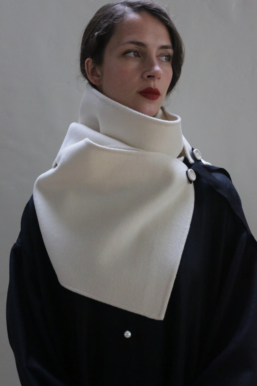 A person with fair skin and brown hair is wearing a stylish black coat and a cream-colored X Scarf - Double-face Cashmere | Undyed. The scarf, made from double-faced cashmere, is wrapped around the neck and fastened with ceramic buttons. The person has a neutral expression and is gazing to the side. The background is plain and neutral-colored.