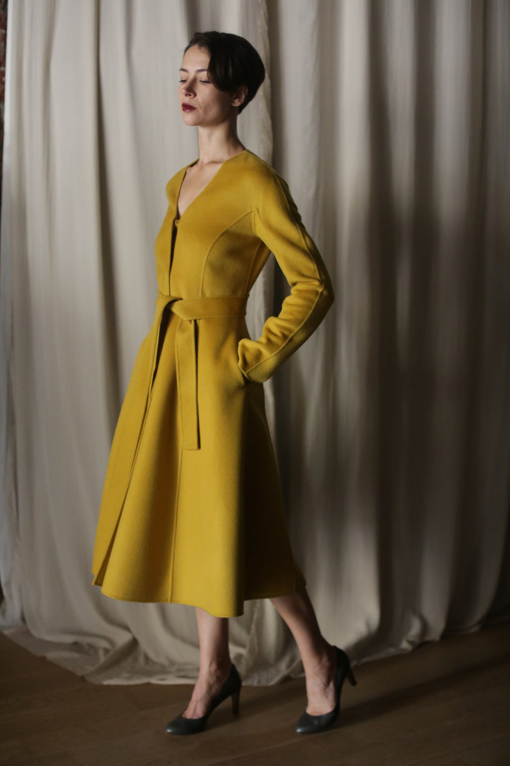 In a softly lit room with beige curtains, a person stands confidently in The Ballerina Coat, a doubleface cashmere piece in saffron. They pose with one foot forward, hands in pockets, complemented by luxurious gray high-heeled shoes.