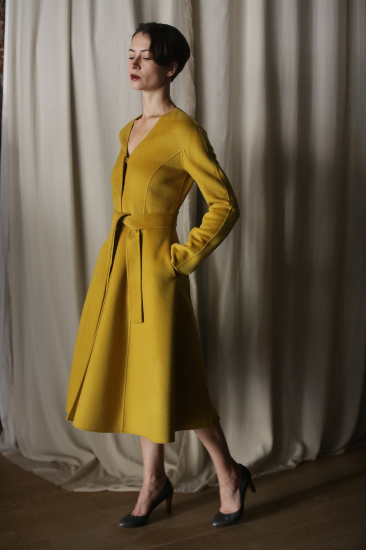 In a softly lit room with beige curtains, a person stands confidently in The Ballerina Coat, a doubleface cashmere piece in saffron. They pose with one foot forward, hands in pockets, complemented by luxurious gray high-heeled shoes.