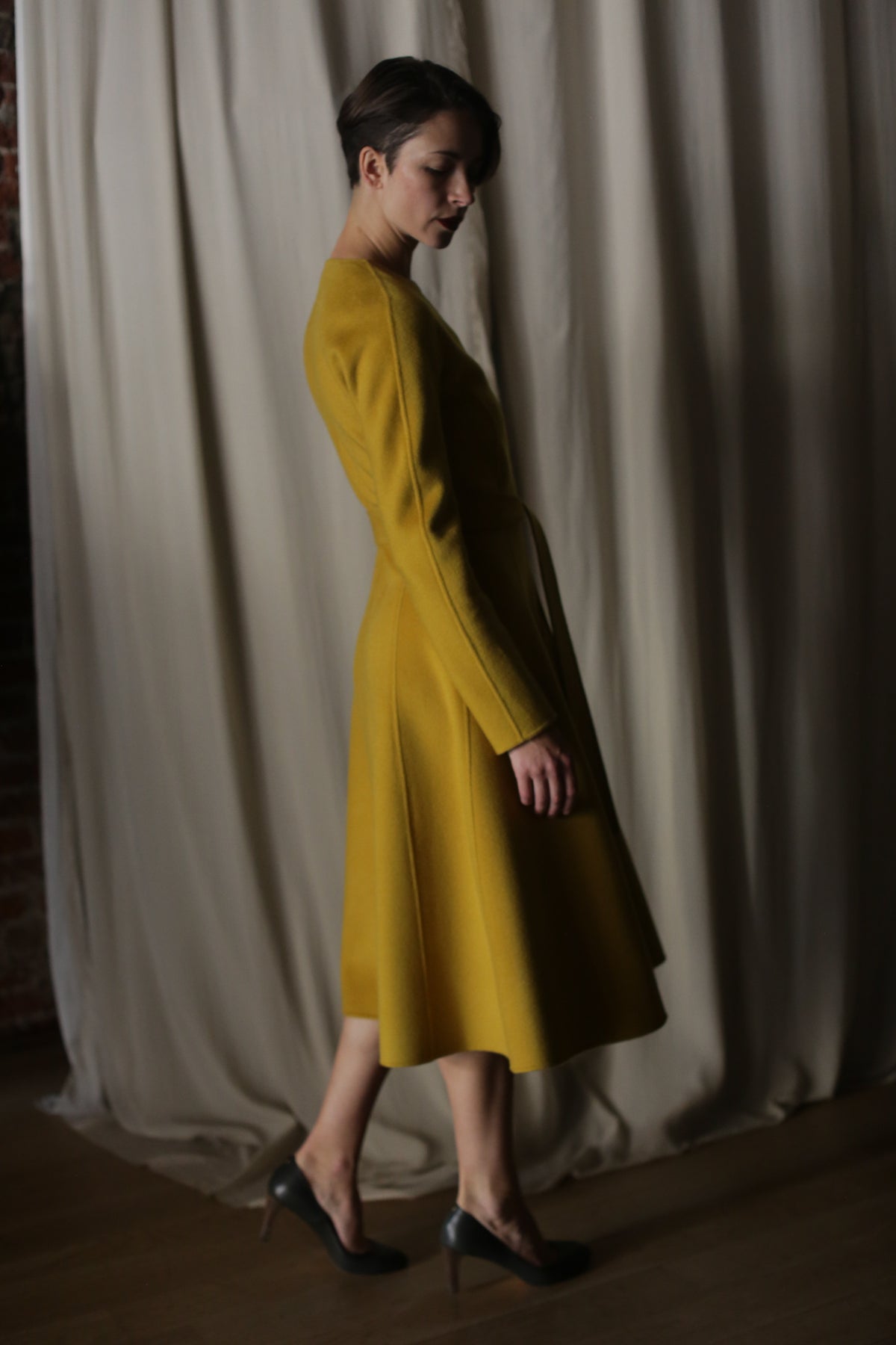 A person wearing The Ballerina Coat in Doubleface Cashmere Saffron and dark heels stands sideways in a dimly lit room with cream curtains, highlighting the coat’s elegant length and luxurious silhouette.