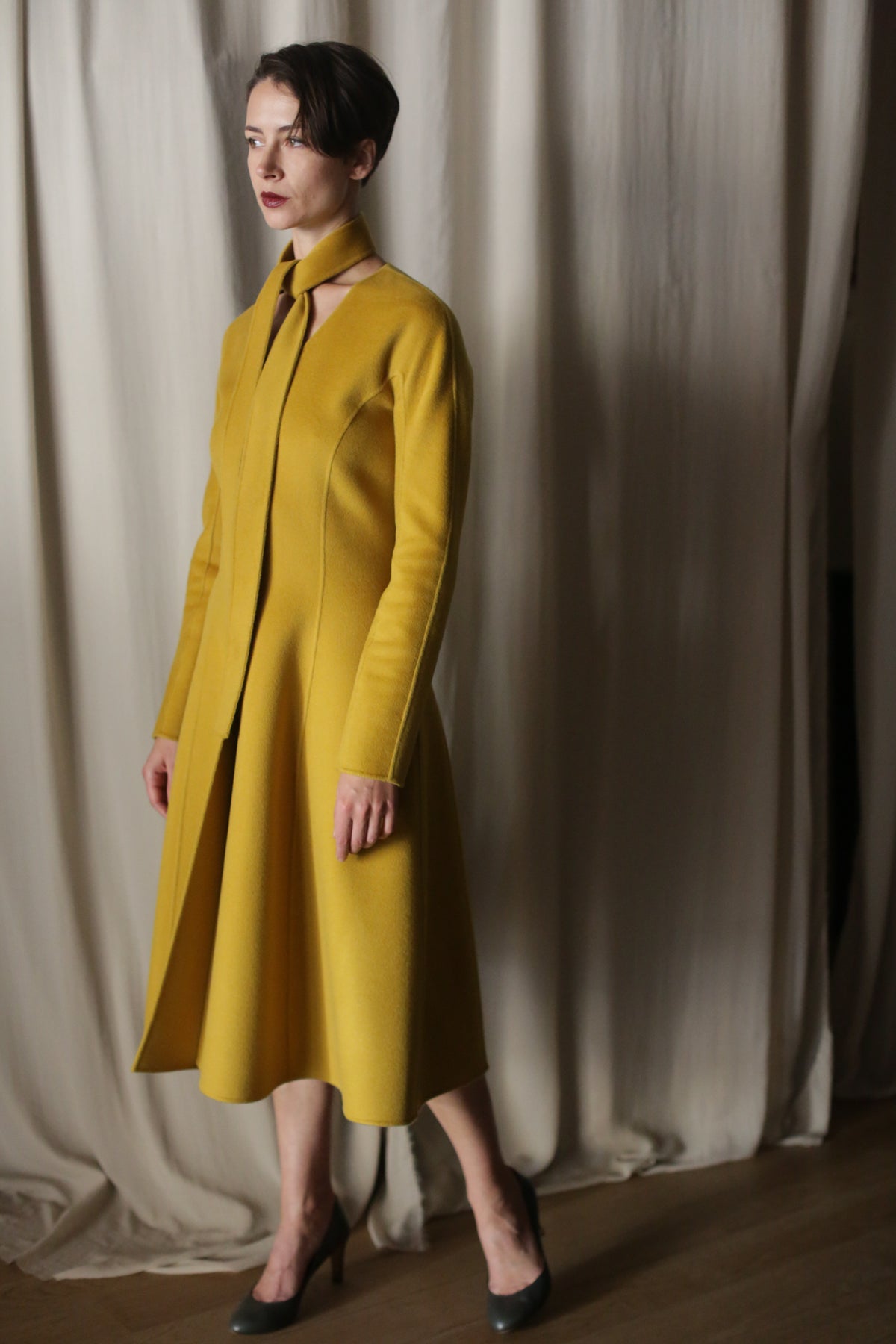 A person stands in front of a curtain wearing The Ballerina Coat in Doubleface Cashmere Saffron, showcasing a unique neckline and tie detail. They pair it with dark heels and their short hair complements their contemplative expression.