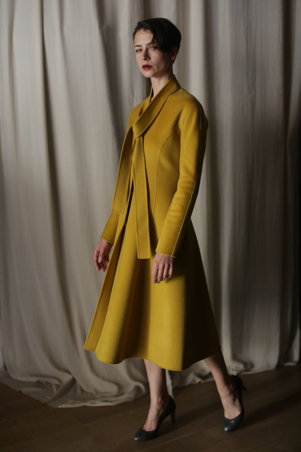 Against luxurious, flowing curtains, a person exudes elegance in a Saffron Doubleface Cashmere Ballerina Coat with a matching skirt and high heels. Soft lighting casts shadows on the textured fabric, highlighting the outfit's shape and color.