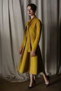 Against luxurious, flowing curtains, a person exudes elegance in a Saffron Doubleface Cashmere Ballerina Coat with a matching skirt and high heels. Soft lighting casts shadows on the textured fabric, highlighting the outfit's shape and color.