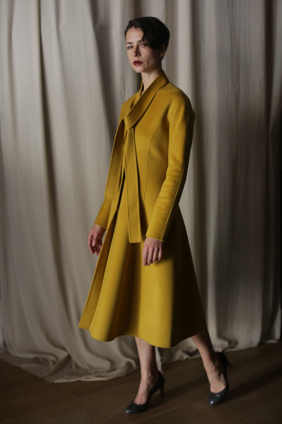 Against luxurious, flowing curtains, a person exudes elegance in a Saffron Doubleface Cashmere Ballerina Coat with a matching skirt and high heels. Soft lighting casts shadows on the textured fabric, highlighting the outfit's shape and color.