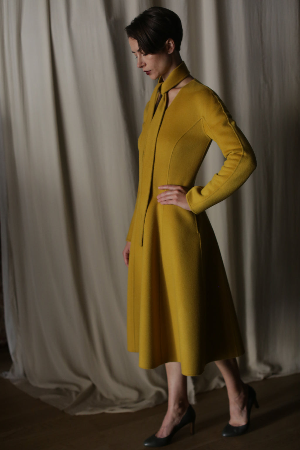 A person stands in profile, radiating luxury in The Ballerina Coat | Doubleface Cashmere Saffron, its mustard-yellow hue echoing elegance. With a high collar and unique tie, they pose confidently with one hand on their hip against a draped curtain backdrop.