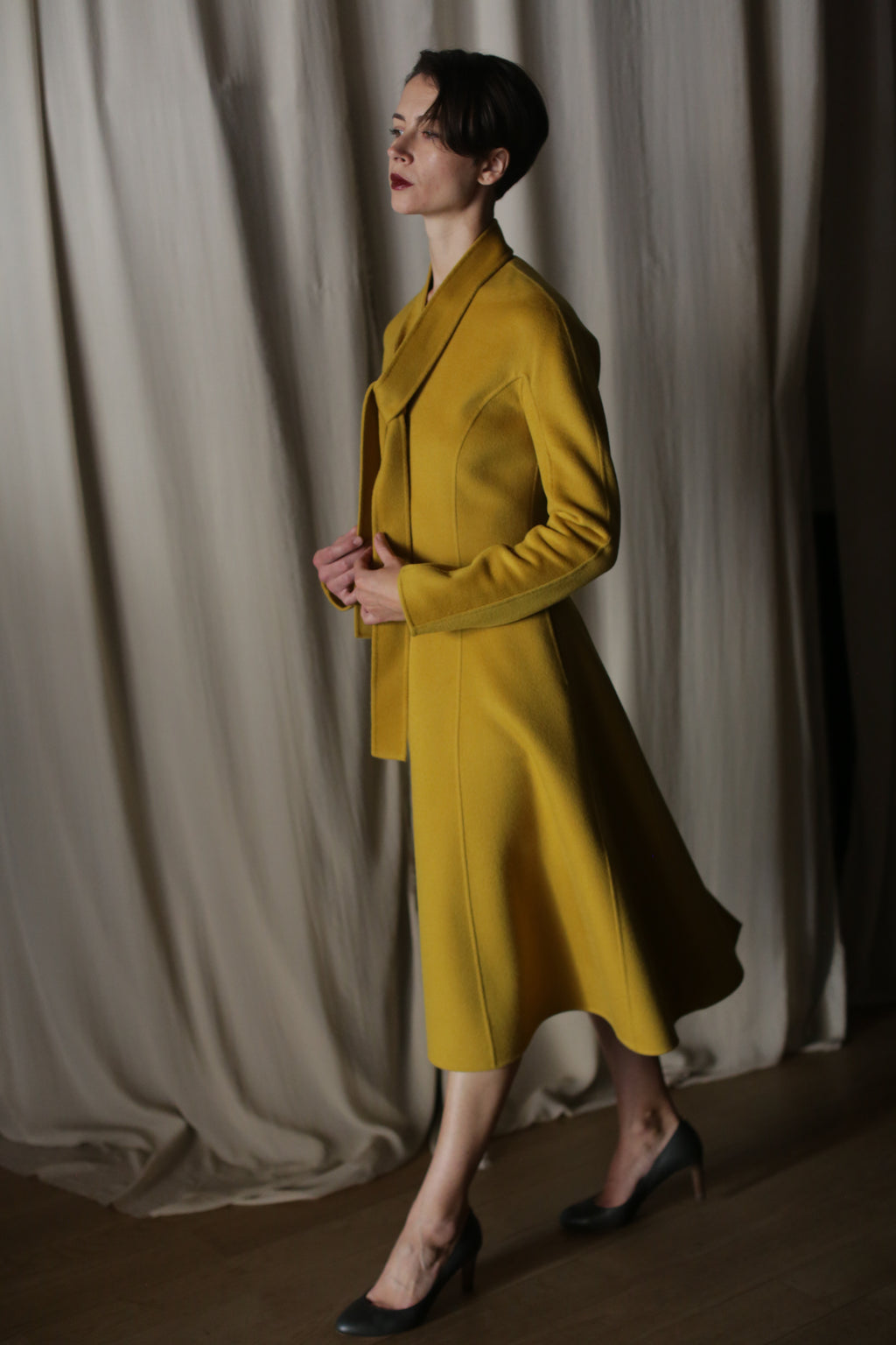 A person in a luxurious The Ballerina Coat | Doubleface Cashmere Saffron stands against a draped curtain background, exuding poise as they gaze to the side with hands gently clasped, complemented by dark heels.