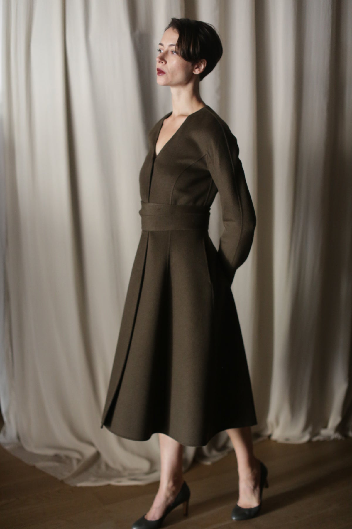 A woman in an olive, doubleface cashmere Ballerina Coat with long sleeves stands by a draped curtain. She has short hair, wears high heels, and looks to the side, arms behind her back.
