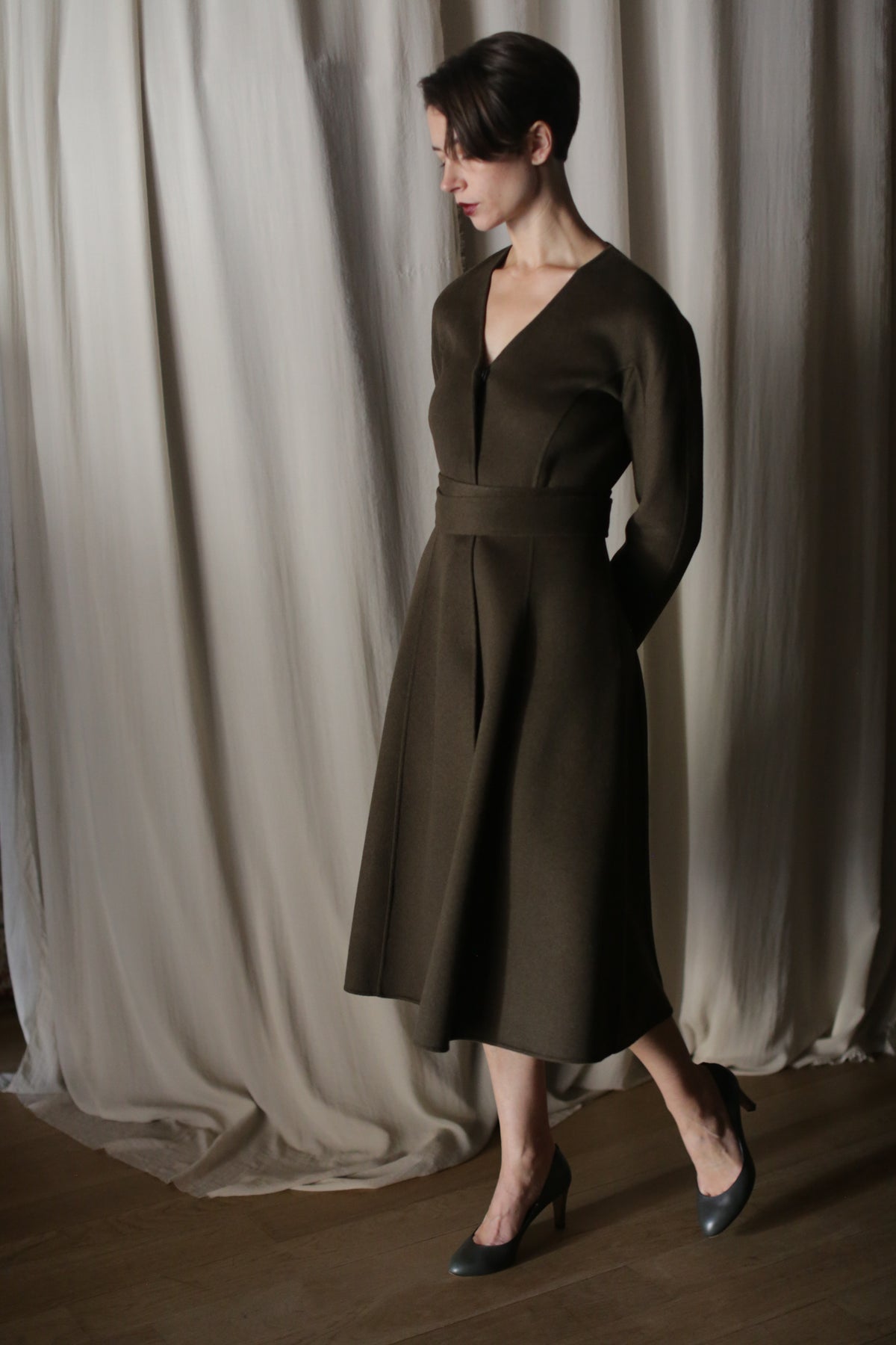 A person wears The Ballerina Coat in Doubleface Cashmere Olive with its elegant silhouette, standing on a wooden floor in black heels. They have short, dark hair, set against a backdrop of draped light-colored fabric.