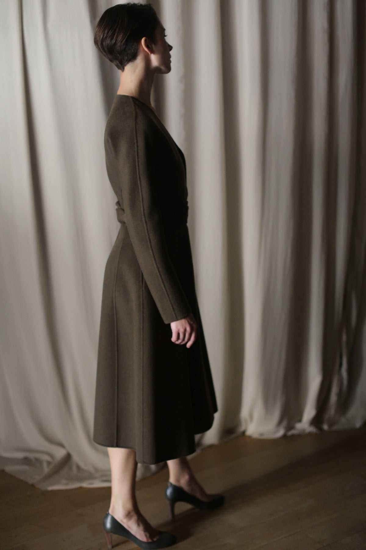 Against light-colored drapes, a person stands in profile, wearing The Ballerina Coat. This luxury coat, featuring a tailored silhouette and crafted from olive double-faced cashmere, pairs elegantly with dark shoes as gentle lighting casts soft shadows.