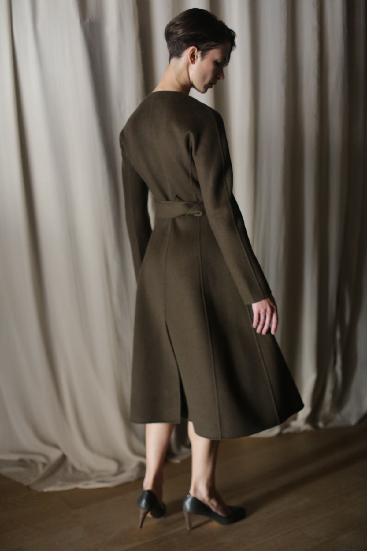 Wearing "The Ballerina Coat | Doubleface Cashmere Olive," a person showcases its luxurious, tailored silhouette with long sleeves and a back tie. Their short hair and high heels complement the look, set against a curtained background.