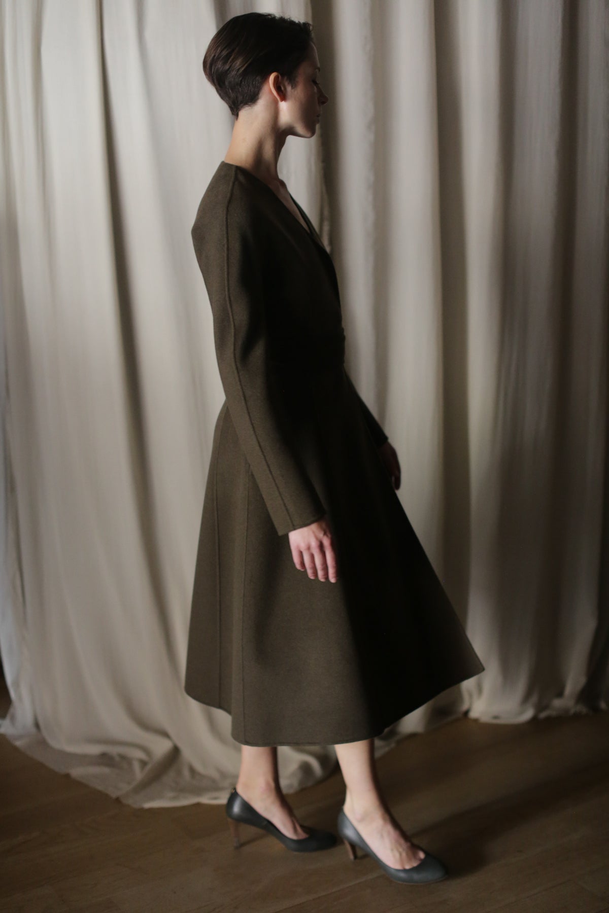 Wearing The Ballerina Coat in double-faced cashmere olive and high heels, a person stands in profile against softly draped curtains, enhancing the tailored silhouette and dimly elegant ambiance.