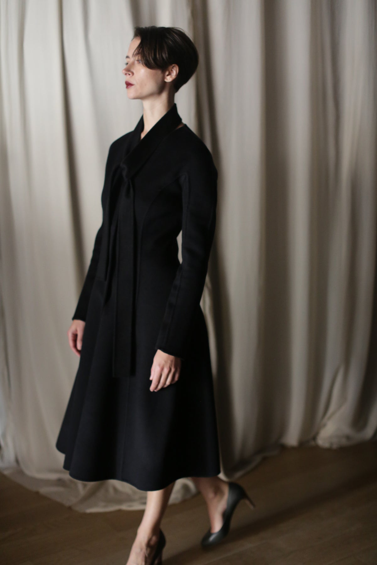 A person in profile wears The Ballerina Coat, a long, black Doubleface Cashmere coat with a tailored silhouette and heels, set against flowing light curtains.
