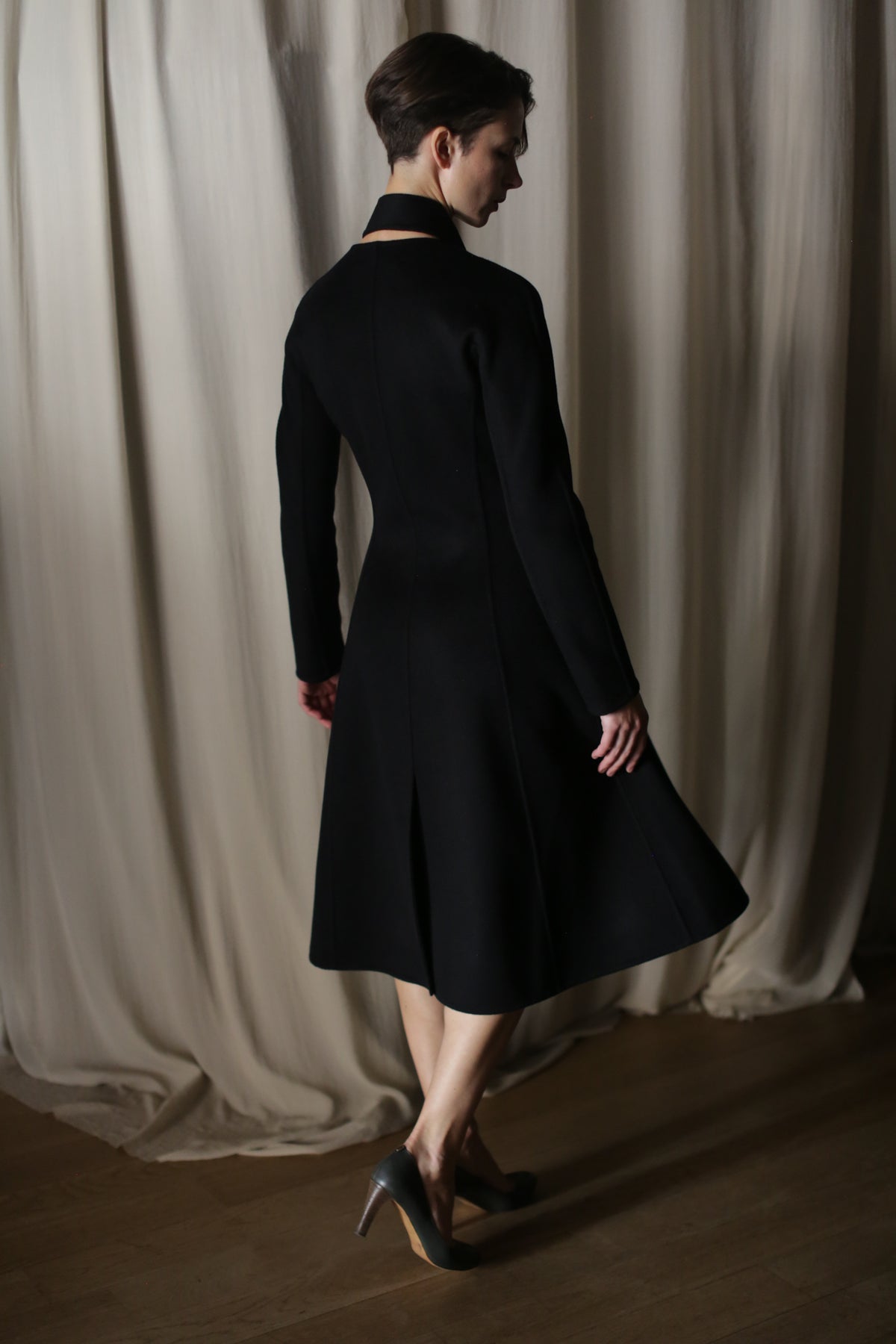 A person wearing The Ballerina Coat in Doubleface Cashmere Black and high heels stands on a wooden floor before light-colored curtains, partially turned away from the camera, highlighting a tailored silhouette that exudes luxury fashion.