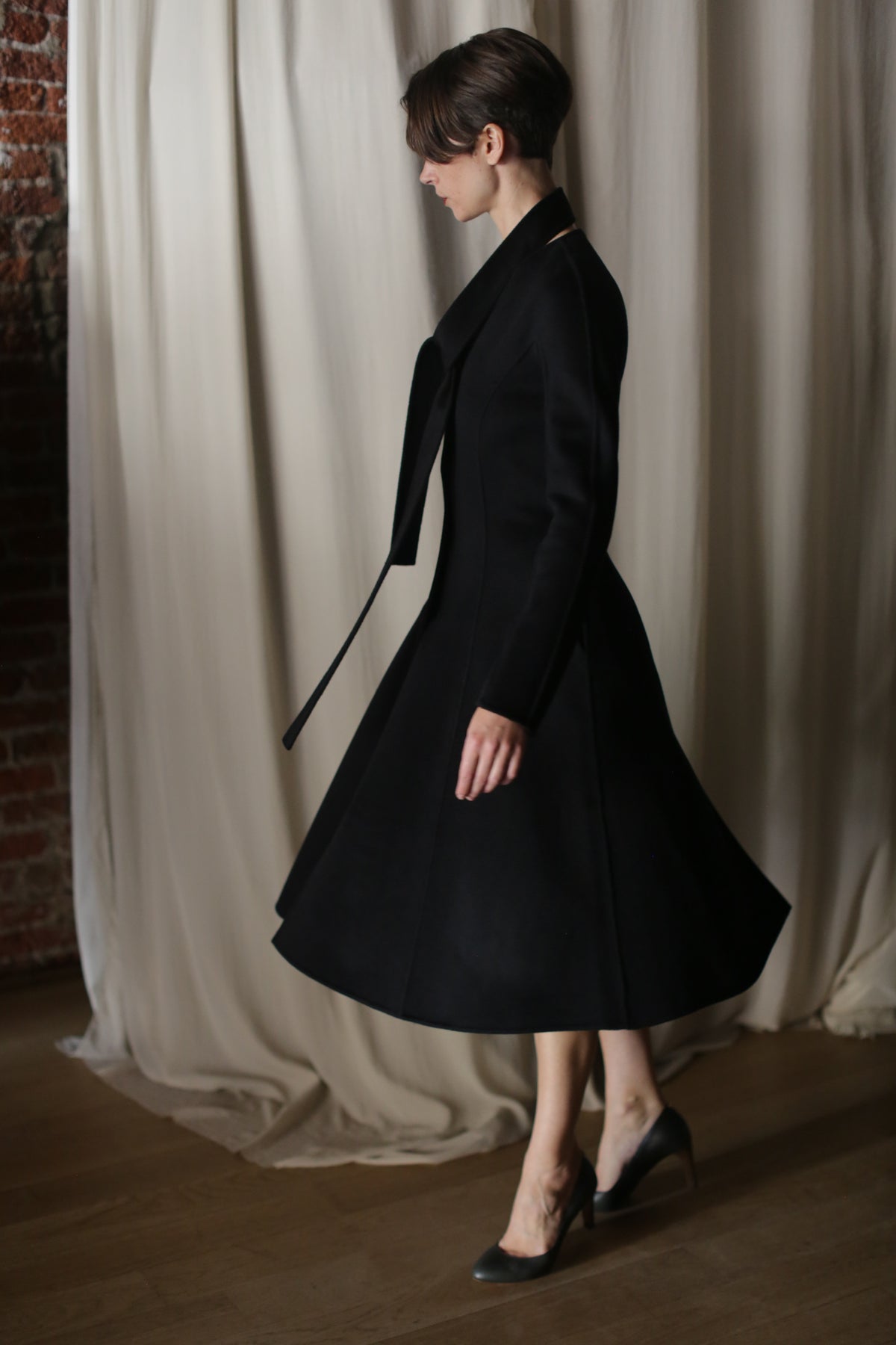 A person wearing a high-heeled luxury fashion ensemble with The Ballerina Coat in Doubleface Cashmere Black is captured in profile, standing before a light-colored curtain alongside a partially visible brick wall.