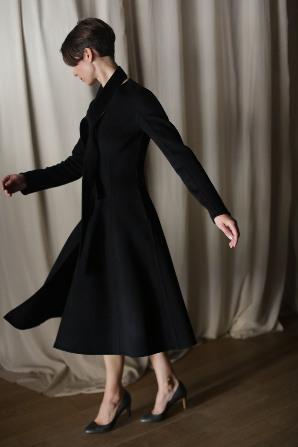 Wearing "The Ballerina Coat" in doubleface black cashmere and high heels, a person elegantly spins on a wooden floor. The tailored silhouette fits perfectly into the luxurious scene, accentuated by softly draped white curtains.