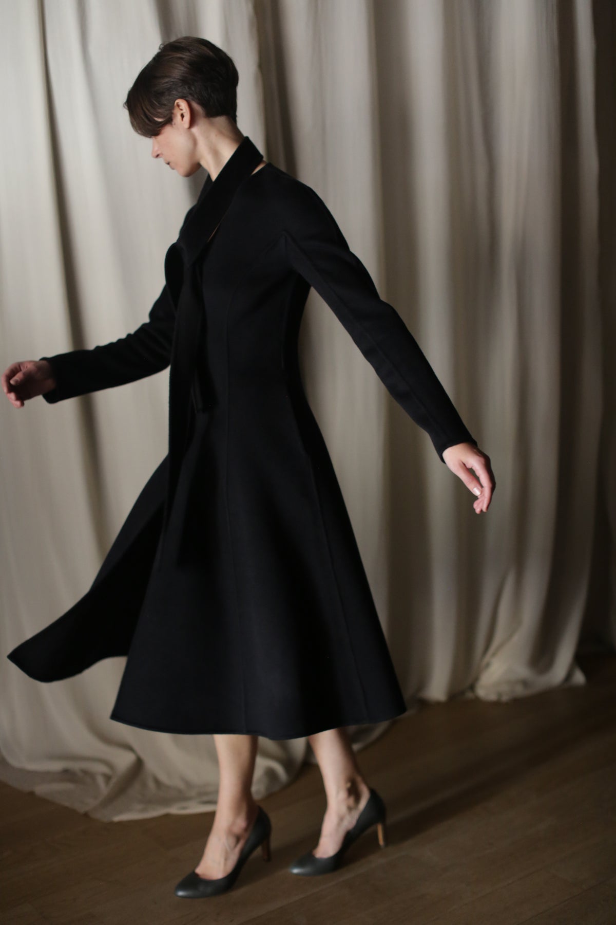 Wearing "The Ballerina Coat" in doubleface black cashmere and high heels, a person elegantly spins on a wooden floor. The tailored silhouette fits perfectly into the luxurious scene, accentuated by softly draped white curtains.