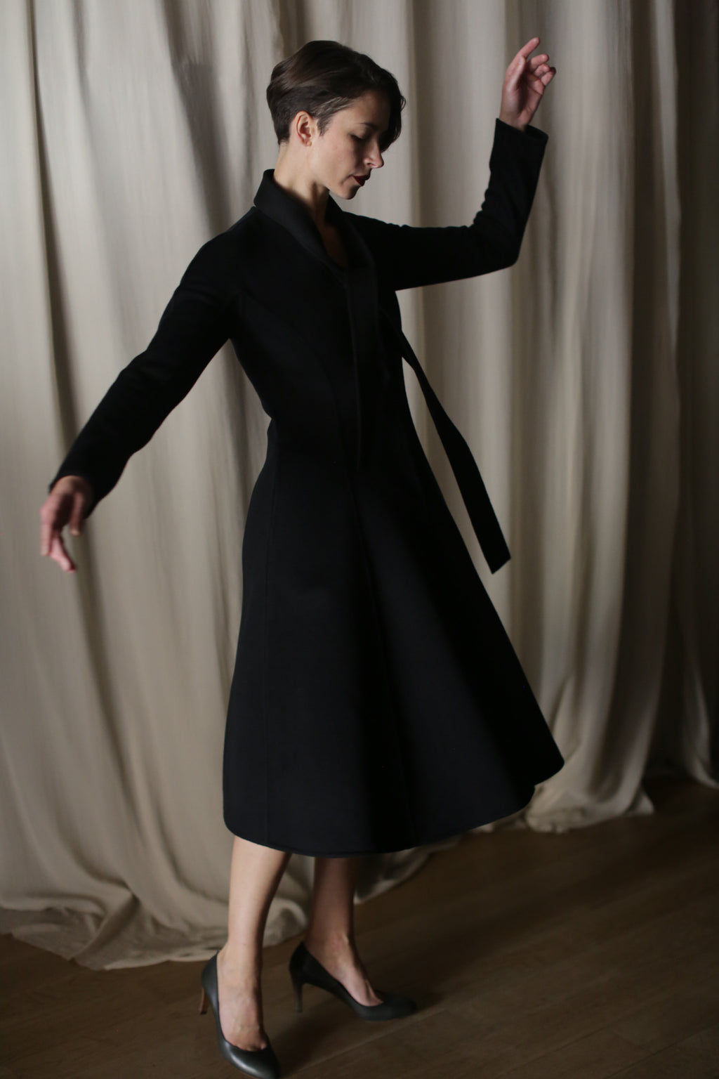 A person wearing The Ballerina Coat in Doubleface Cashmere Black stands gracefully on one foot with arms extended against flowing curtains. Soft lighting highlights the coat's tailored silhouette and elegant lines, complementing the poised posture.