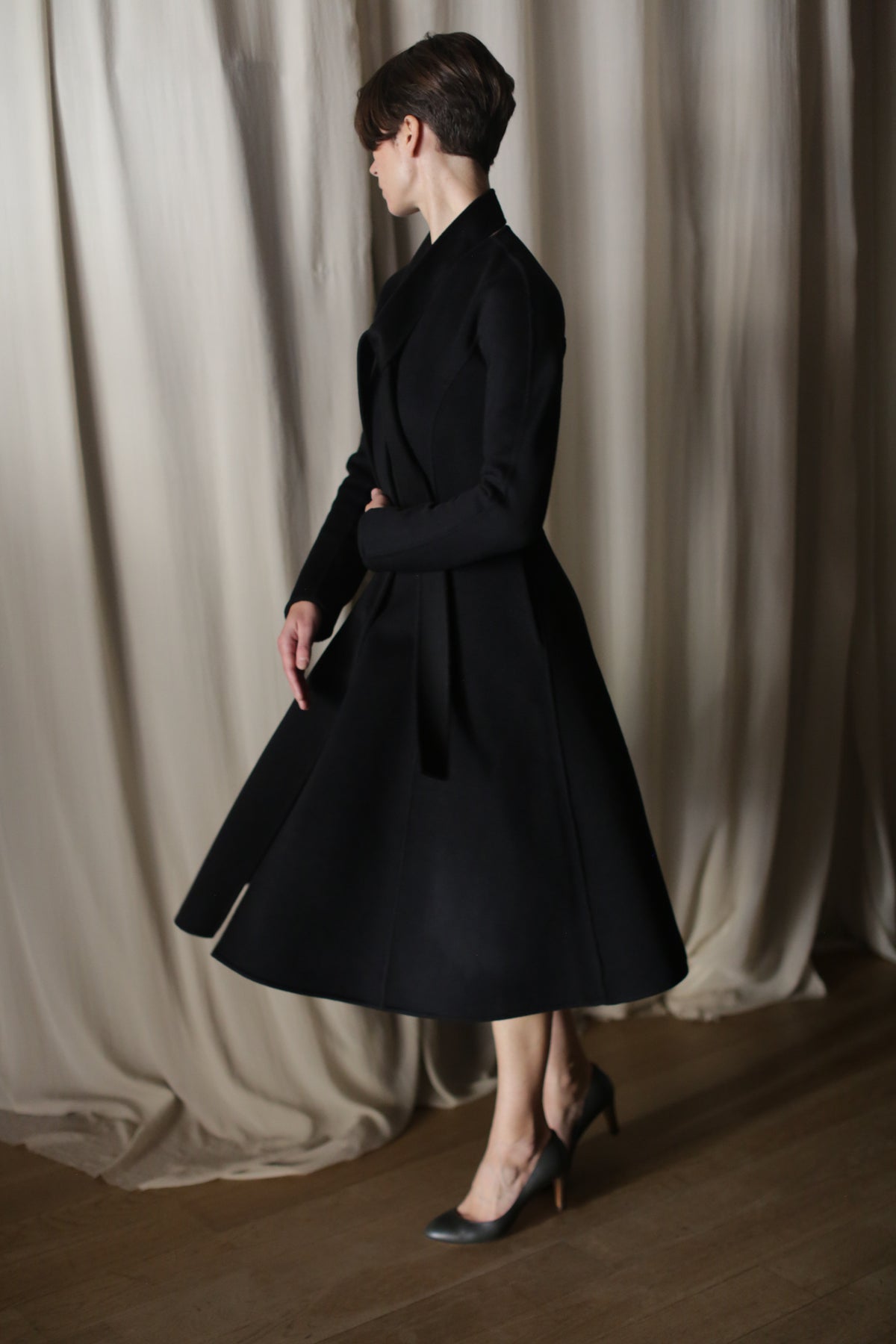 In a dimly lit room with soft, draped curtains, a person elegantly stands sideways with short hair, showcasing The Ballerina Coat in black Doubleface Cashmere and gray high-heeled shoes, epitomizing luxury fashion with its tailored silhouette and flowing design.
