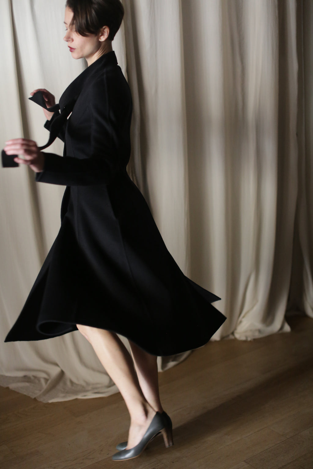 In front of beige curtains, a woman gracefully spins in "The Ballerina Coat | Doubleface Cashmere Black," exuding elegance with her short hair and stylish outfit. The blurred motion captures her fluid movement, while the wooden floor adds warmth to this luxurious fashion scene.