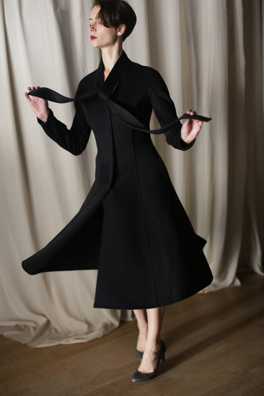 A person in "The Ballerina Coat | Doubleface Cashmere Black" stands on a wooden floor with light curtains behind them, holding the collar's strap. The tailored silhouette flares slightly, paired with high heels.