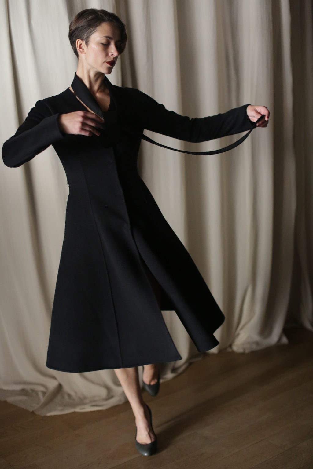 A person twirls gracefully on a wooden floor against an off-white curtain backdrop, wearing The Ballerina Coat in Doubleface Cashmere Black. The tailored silhouette swings outward as they spin, complemented by sleek black shoes with short heels.