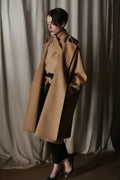 A person stands indoors against cream curtains, wearing the Kyoto | Camel luxury double-faced cashmere coat draped over their shoulders, paired with a matching turtleneck, black pants, and heels. Hands in their coat pockets, they look down thoughtfully.