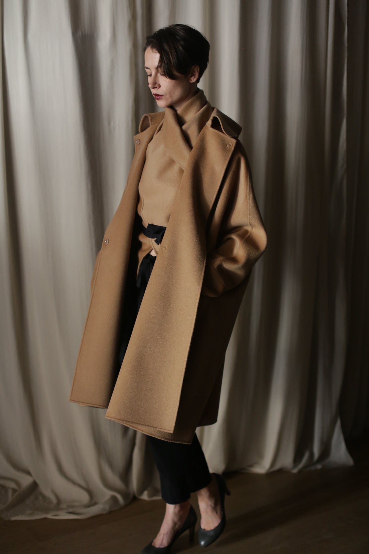 A person stands indoors against cream curtains, wearing the Kyoto | Camel luxury double-faced cashmere coat draped over their shoulders, paired with a matching turtleneck, black pants, and heels. Hands in their coat pockets, they look down thoughtfully.