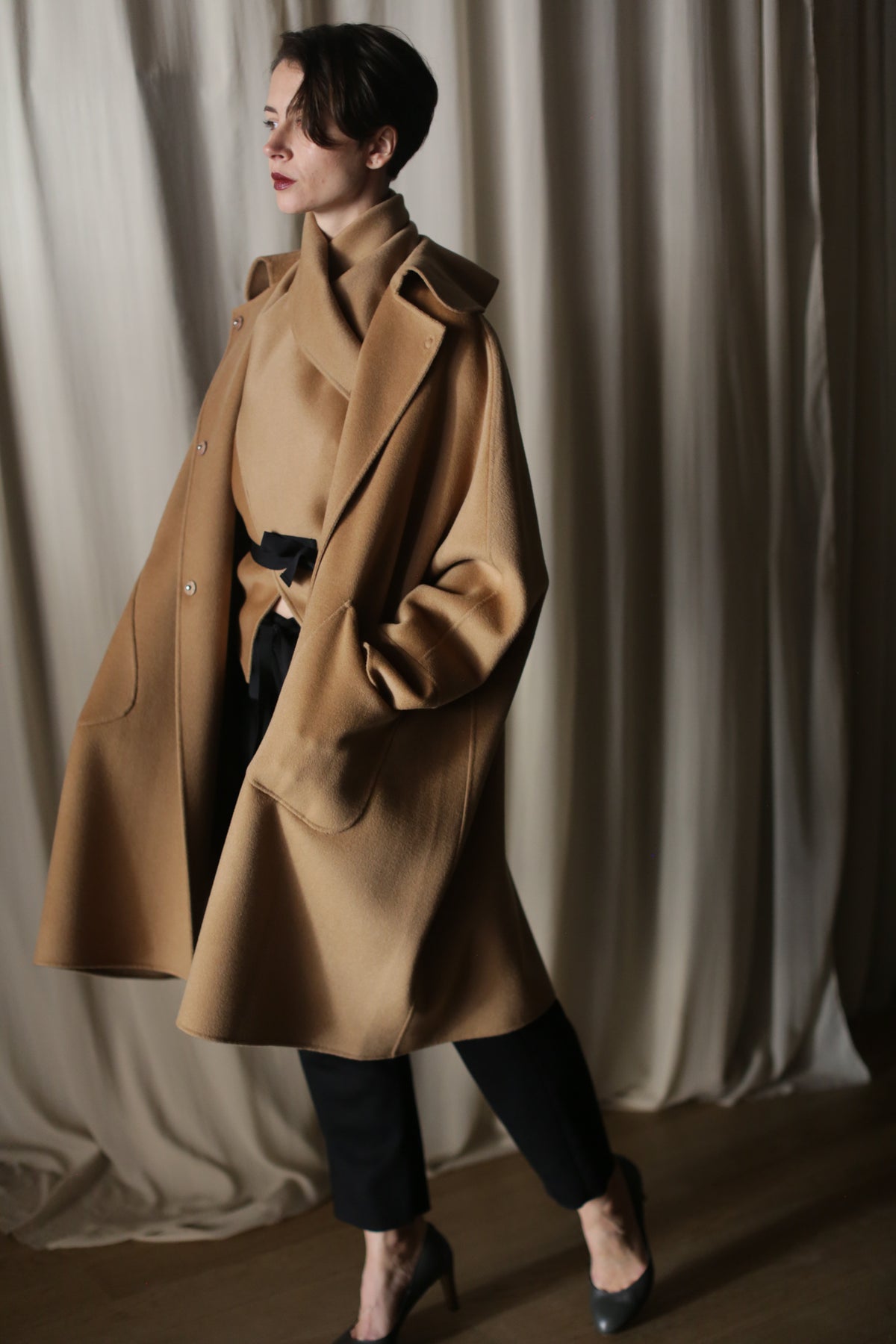 A person stands indoors wearing the Kyoto | Camel luxury women's cashmere coat over a tan turtleneck and black pants, with one hand in their pocket. Cream-colored curtains are in the background.