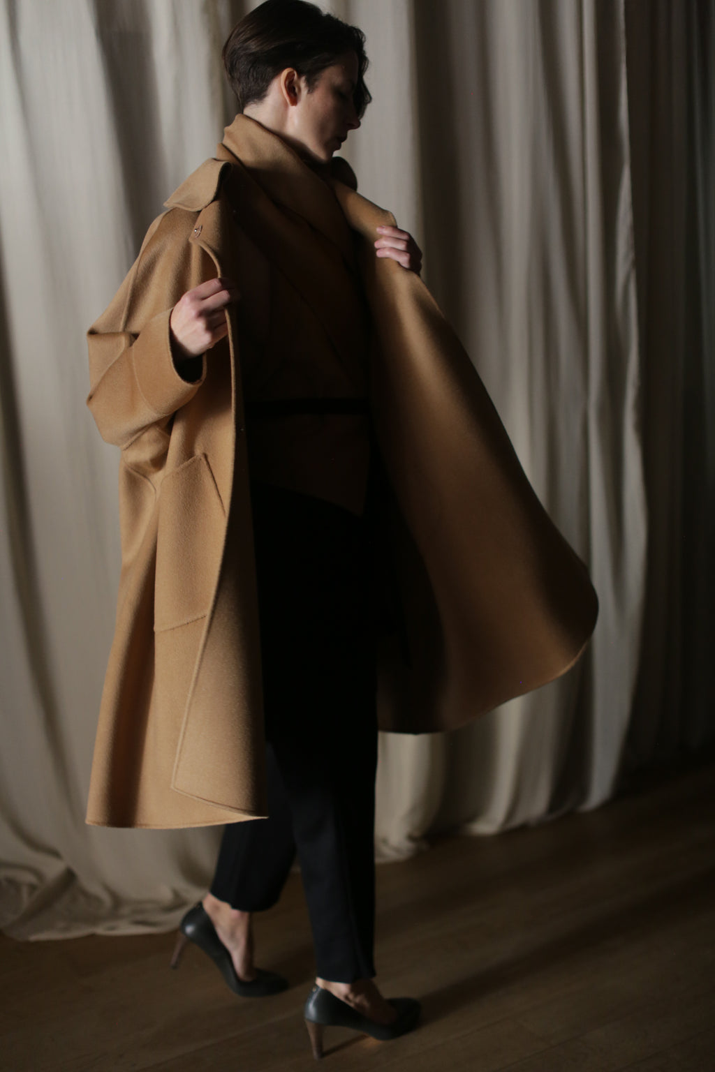Wearing the Kyoto | Camel double-faced cashmere coat and black heels, a person walks indoors on a wooden floor, holding the coat open. Cream-colored curtains are visible in the background.