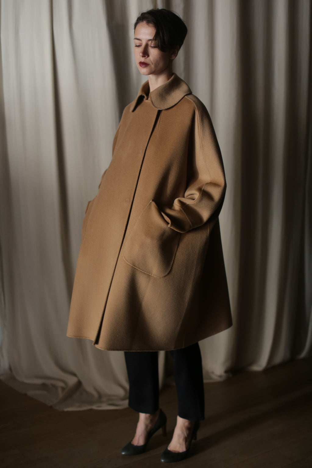 A woman stands indoors before beige curtains, wearing Kyoto | Camel—a loose-fitting luxury cashmere coat with large pockets. She pairs it with black pants and gray heels, her dark hair styled short as she looks down calmly.