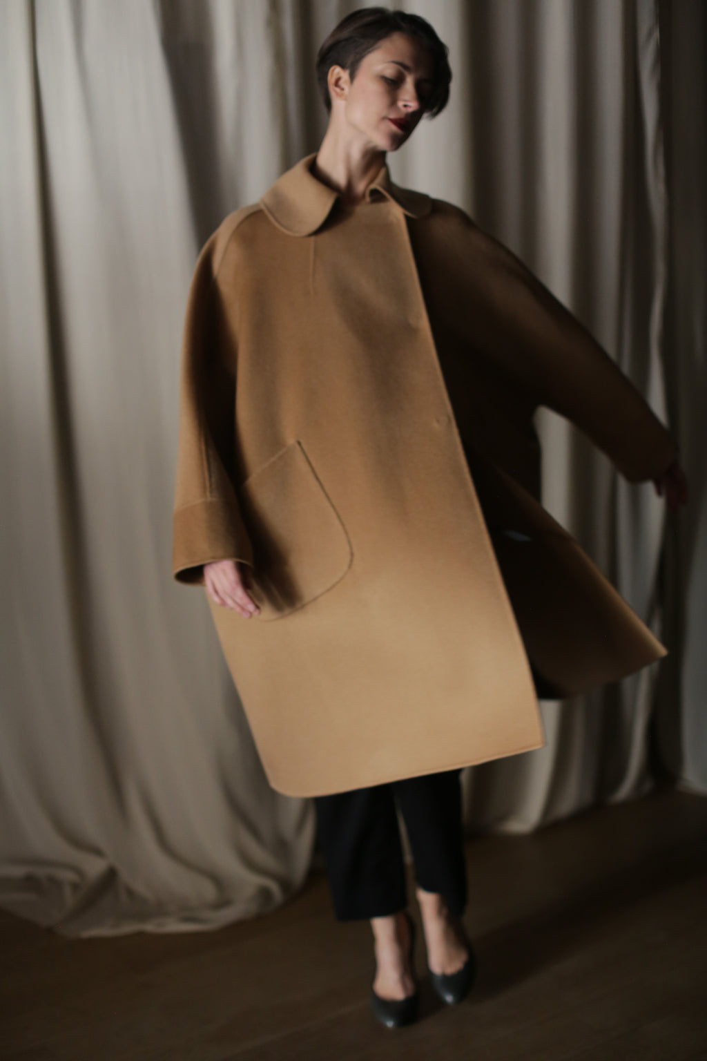 A person stands before cream curtains wearing the Kyoto | Camel coat—a loose, luxury women's cashmere design with wide sleeves and a large pocket—paired with dark pants and heels in soft lighting.