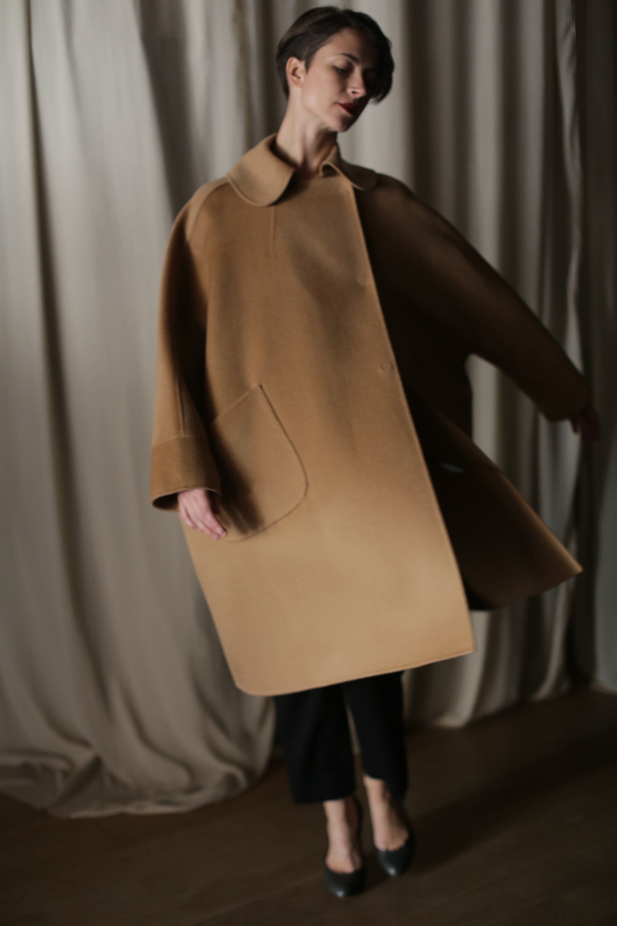 A person stands before cream curtains wearing the Kyoto | Camel coat—a loose, luxury women's cashmere design with wide sleeves and a large pocket—paired with dark pants and heels in soft lighting.