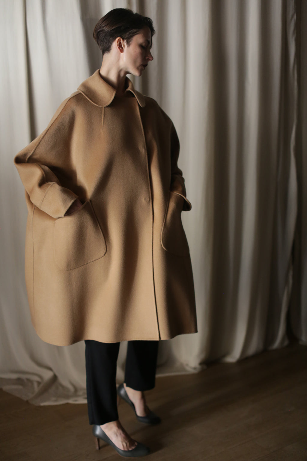 Indoors, a person wears the Kyoto | Camel luxury women's double-faced cashmere coat with large pockets, paired with black pants and heels. Light curtains hang in the background.