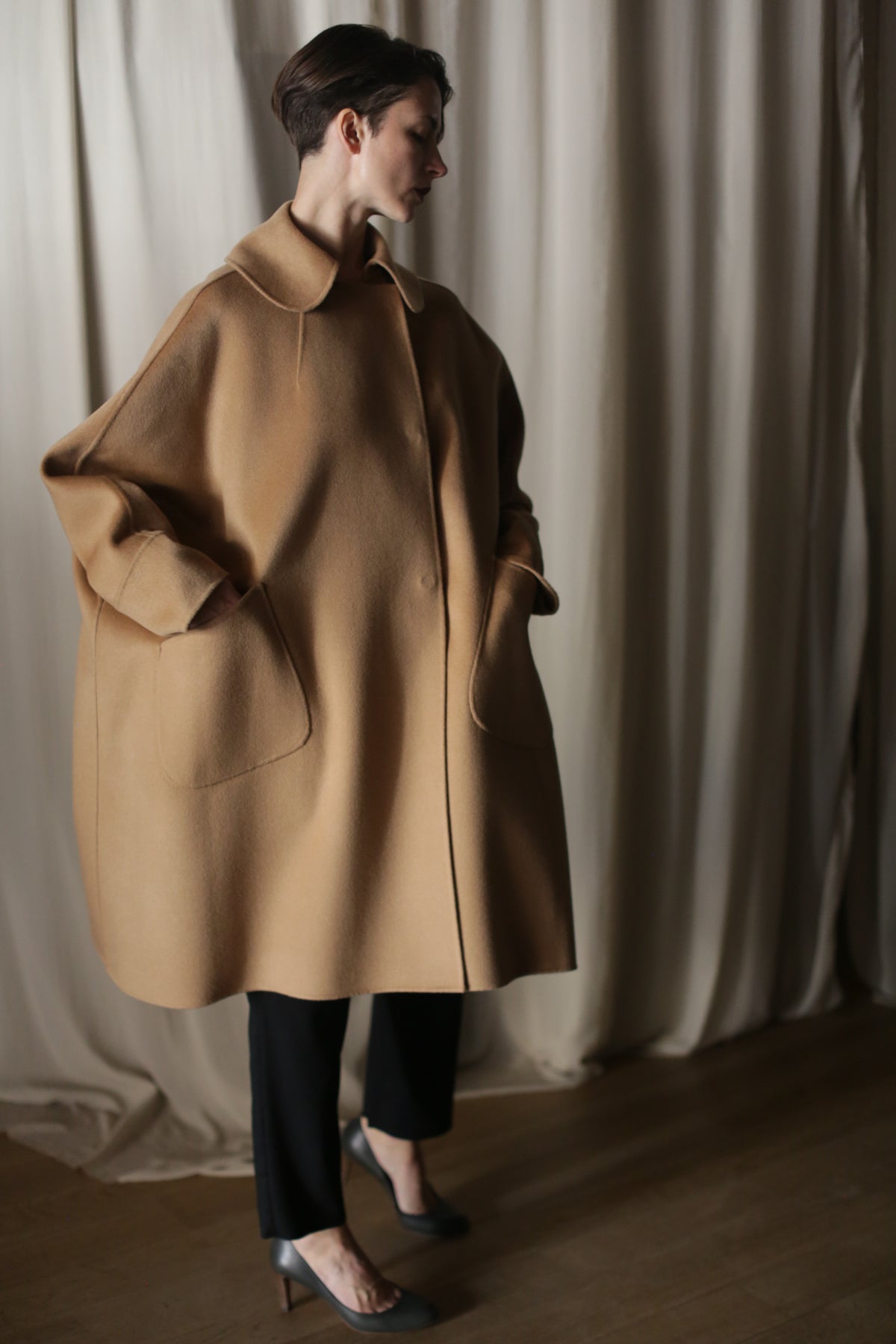 Indoors, a person wears the Kyoto | Camel luxury women's double-faced cashmere coat with large pockets, paired with black pants and heels. Light curtains hang in the background.