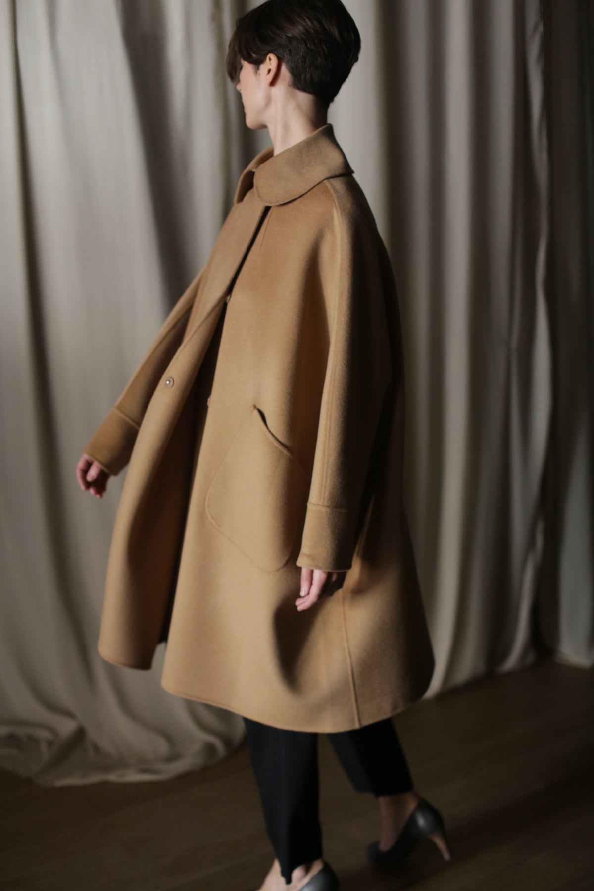 A person with short hair wears the Kyoto | Camel cashmere coat, black pants, and black shoes, standing on a wooden floor before cream-colored curtains with their face turned away from the camera.