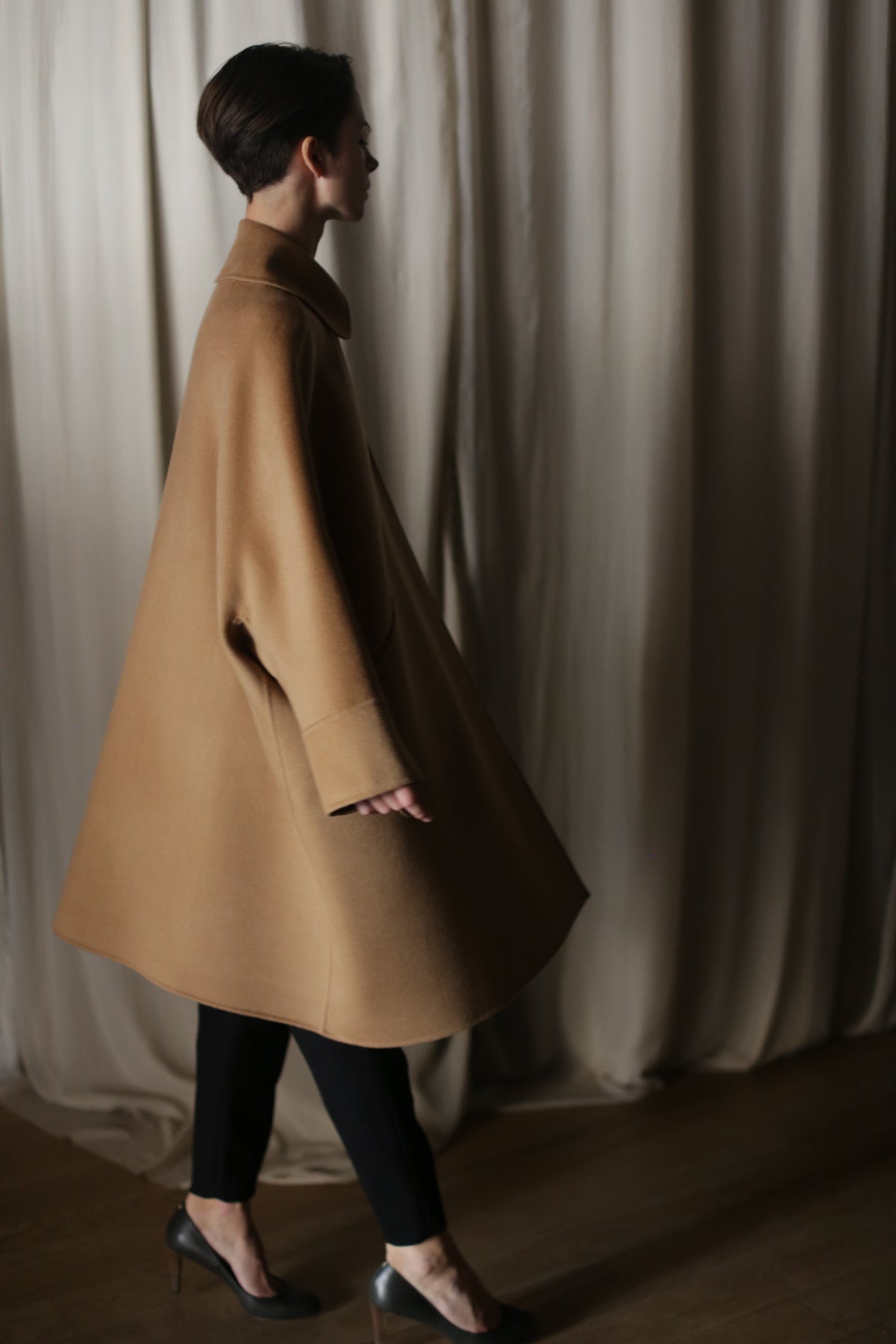 Wearing the Kyoto | Camel luxury women’s double-faced cashmere coat with black pants, a person walks indoors by cream-colored curtains. Soft lighting highlights the coat as she is seen in profile.