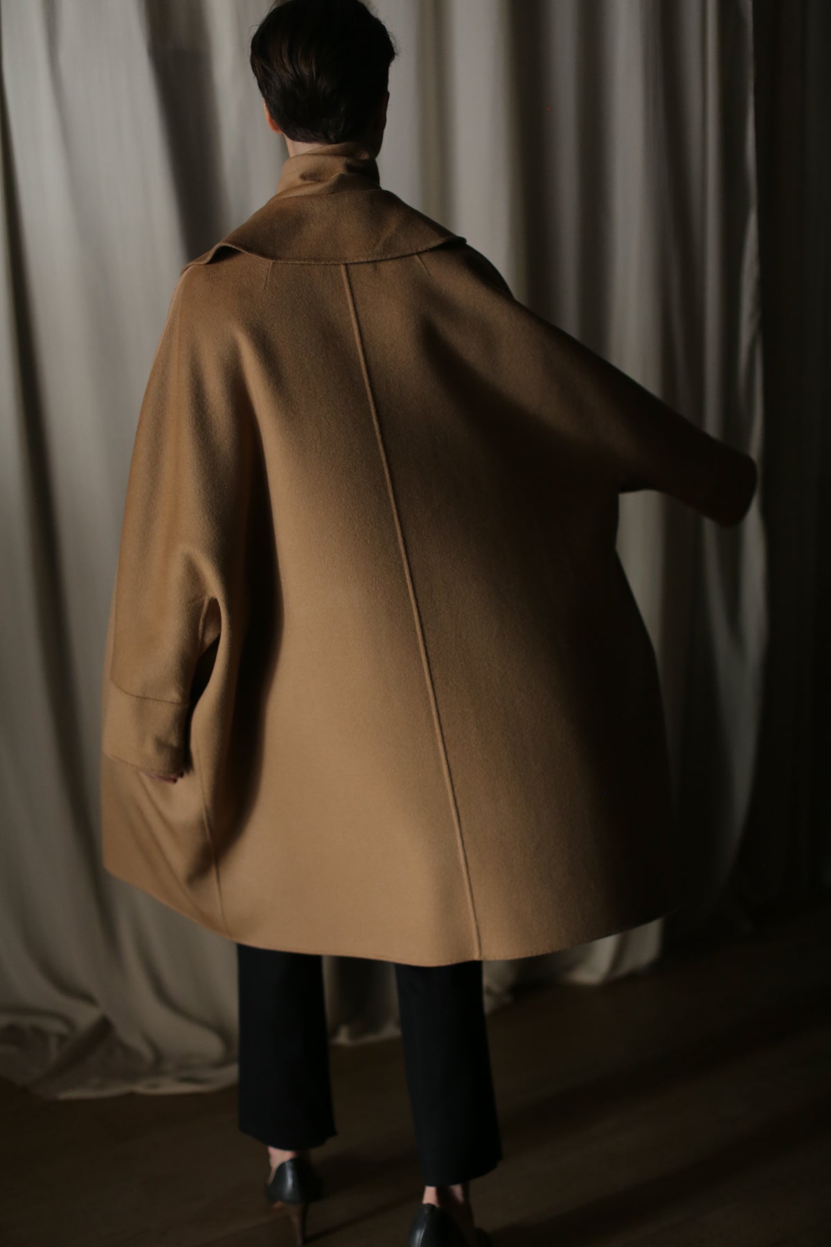 A person stands with their back to the camera, wearing the Kyoto | Camel luxury women's double-faced cashmere coat and black pants, in a dimly lit room with beige curtains in the background.