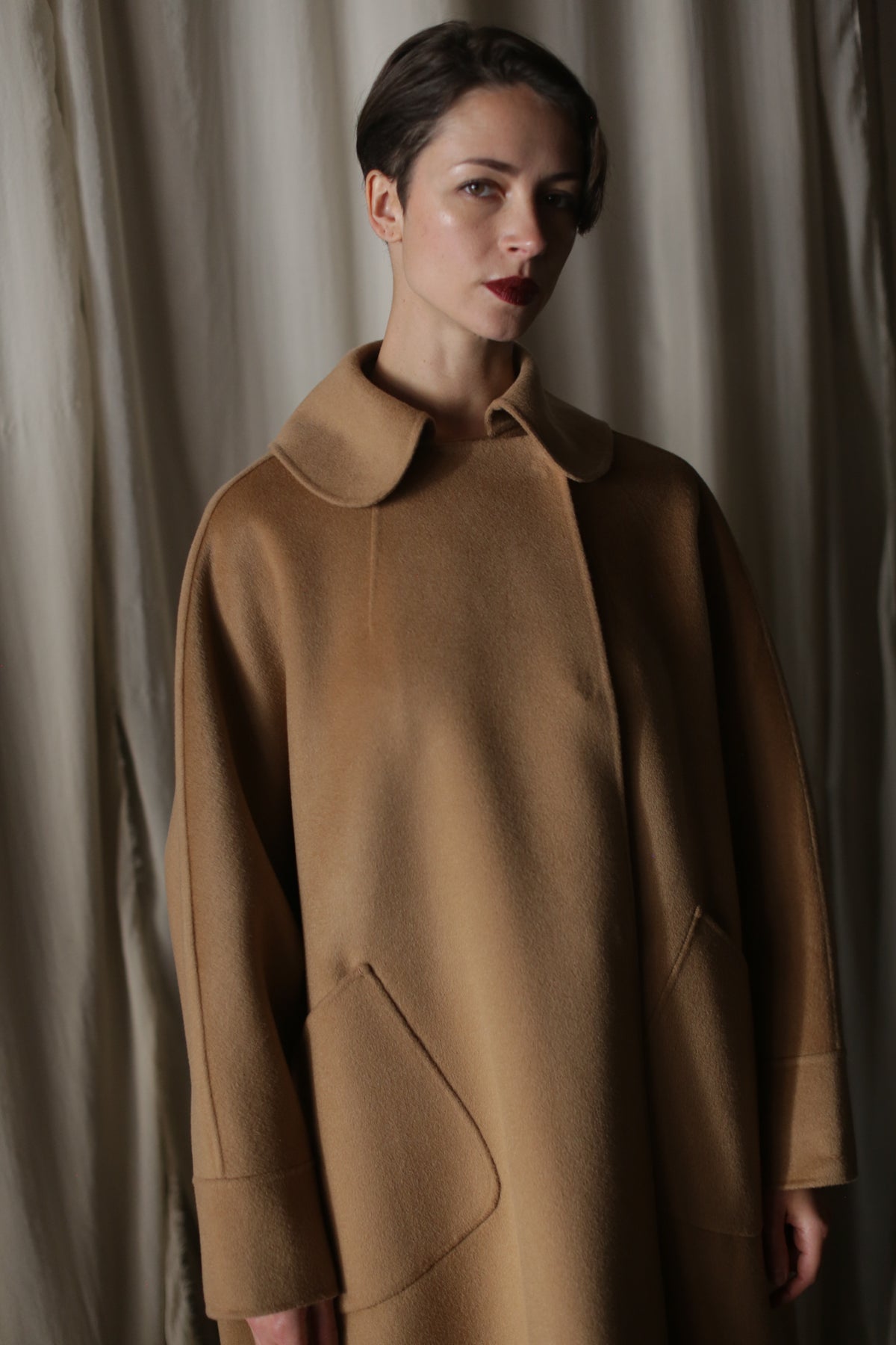 A person with short dark hair wears the Kyoto | Camel luxury women’s coat, standing in front of cream-colored curtains and gazing slightly to the side with a neutral expression.