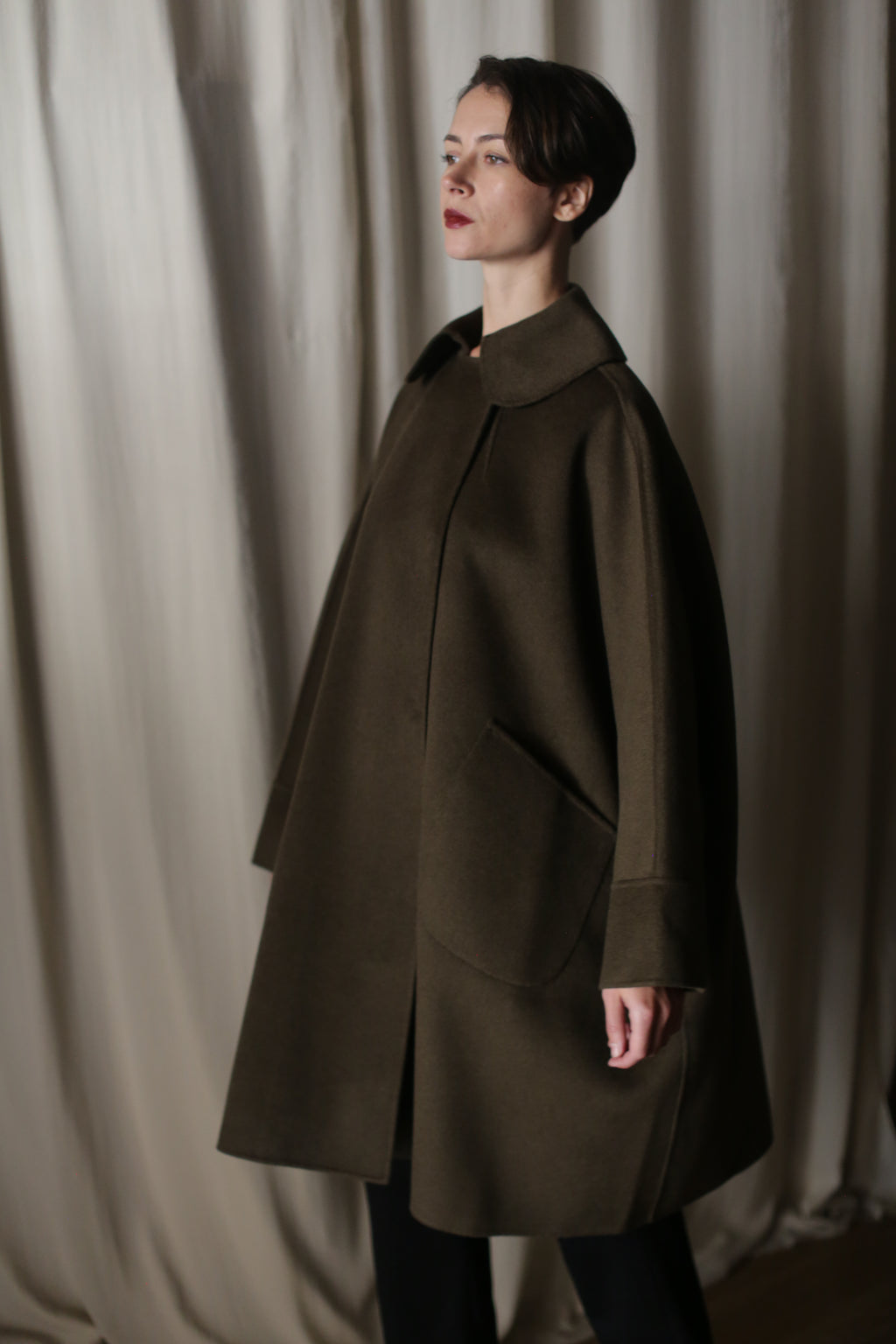 A person with short dark hair stands in front of beige curtains, wearing the oversized "Kyoto | Olive" coat with large pockets. The garment's dramatic silhouette suggests a voluminous shape as they gaze thoughtfully to the side.