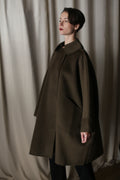 A person with short dark hair stands in front of beige curtains, wearing the oversized "Kyoto | Olive" coat with large pockets. The garment's dramatic silhouette suggests a voluminous shape as they gaze thoughtfully to the side.