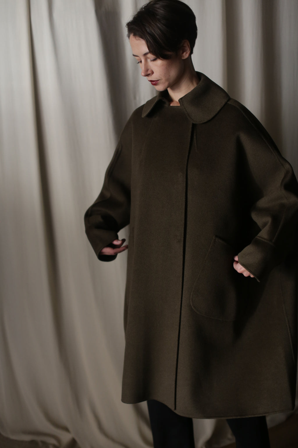 Wearing the Kyoto | Olive coat against a neutral draped background, a person with short dark hair looks down as they tuck their hands into sizable pockets. Crafted from double-faced cashmere, the coat's dramatic silhouette stands out.